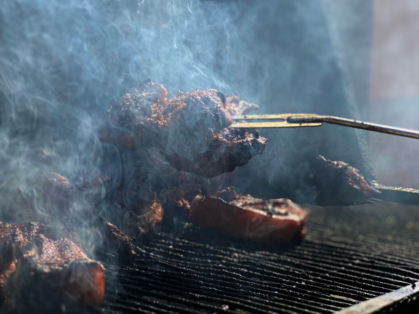 グリル上の肉にトングを添え、白い煙が勢いよく立ちのぼっている様子